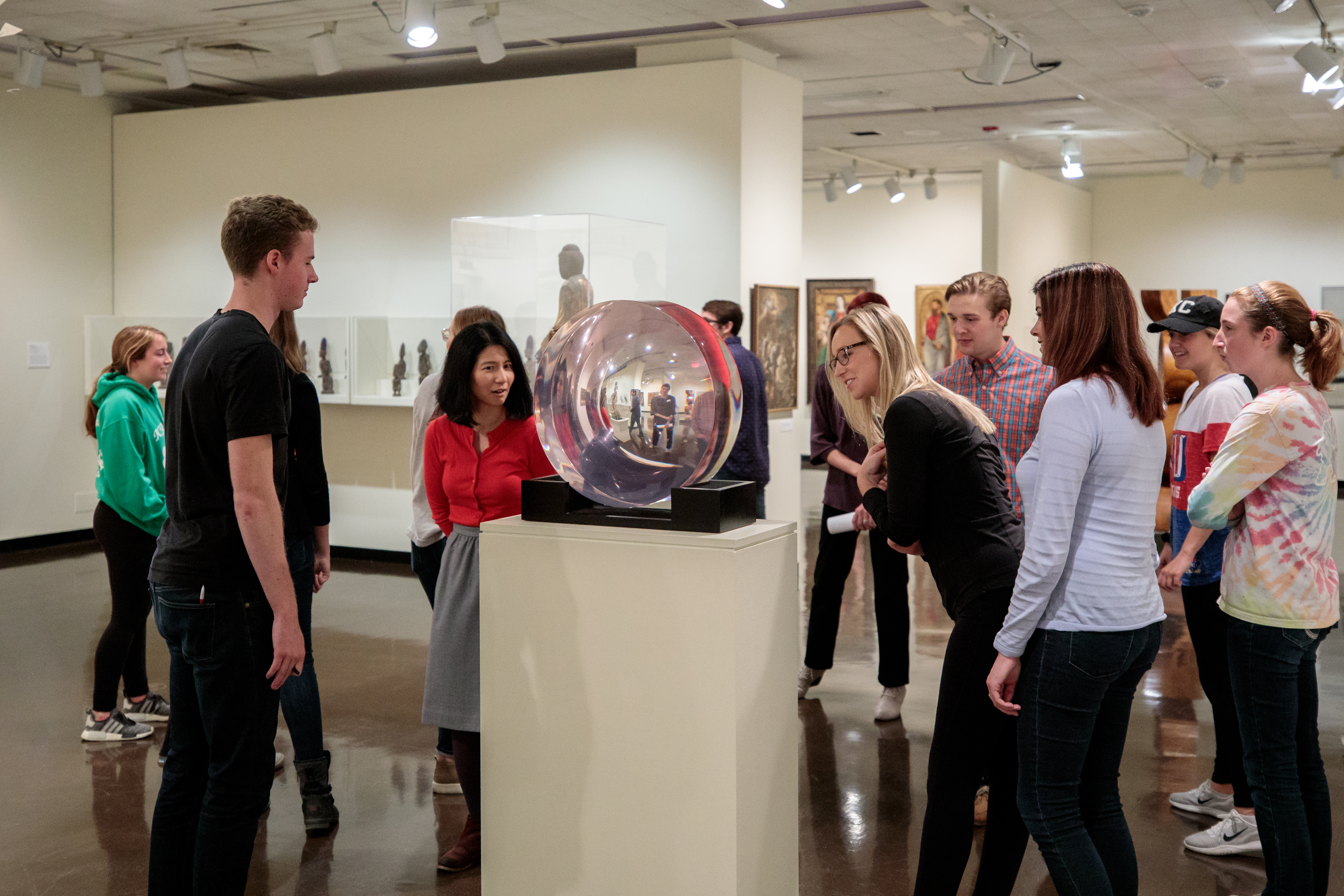 A class gathers around a round glass piece of art that reflects their images