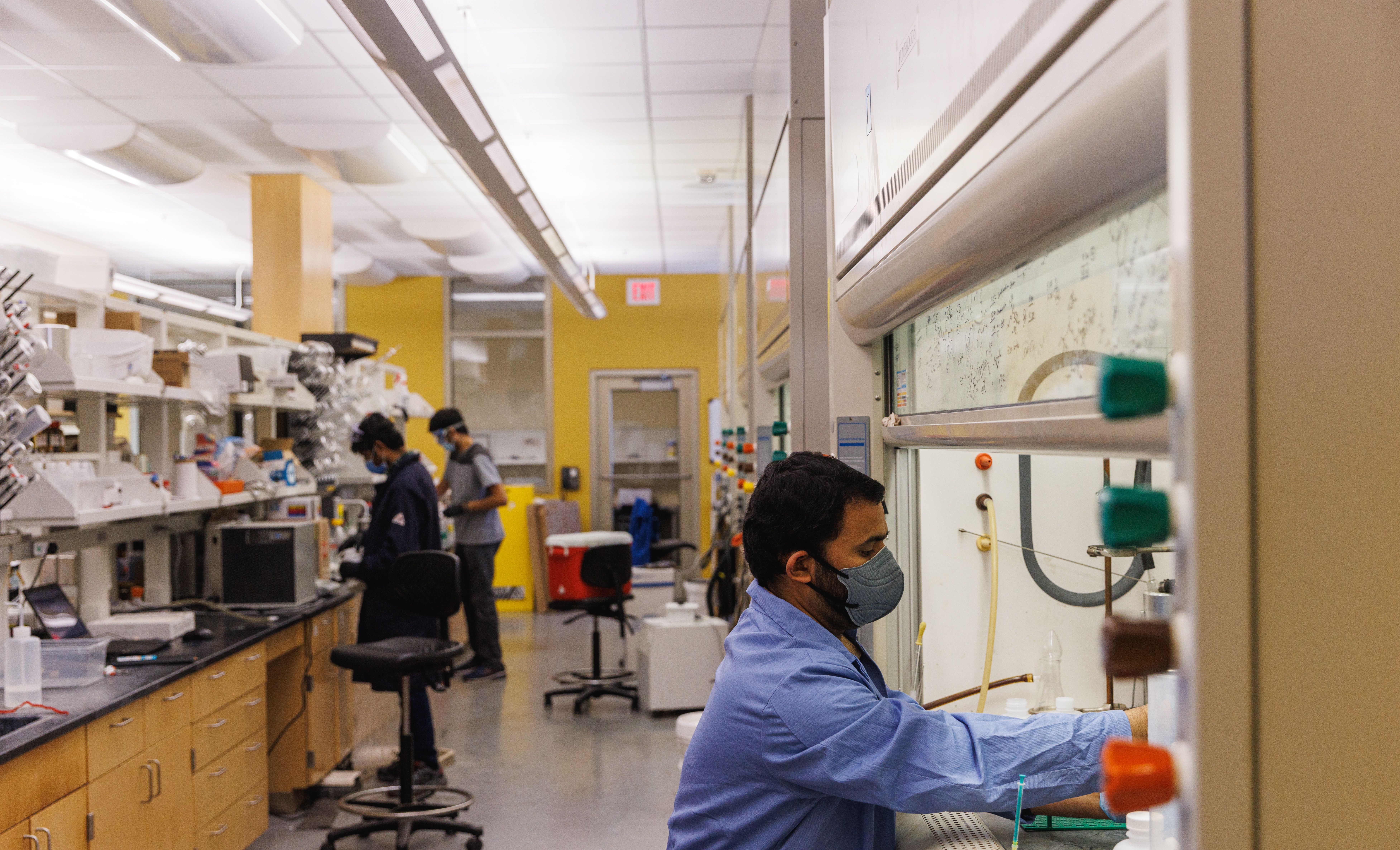 Three people work at different stations in a pharmaceutical chemistry lab