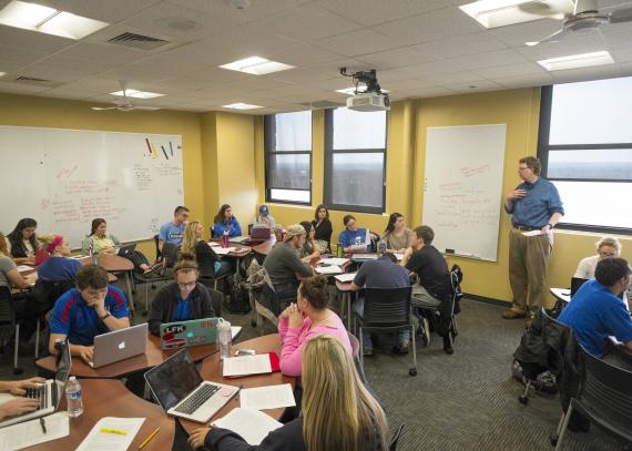 Students at tables and on laptops listen to a lecture in a classroom