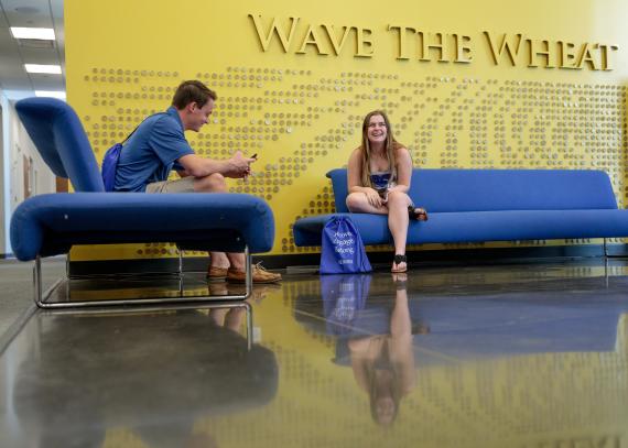 two students sit in front of a wall that says, “Wave the Wheat” in Capitol Federal Hall