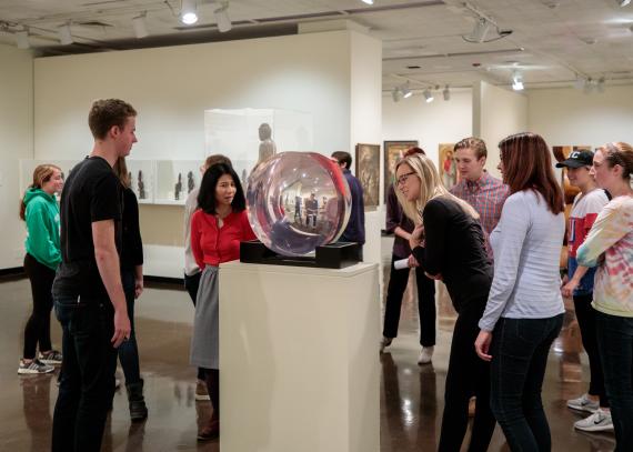 A class gathers around a round glass piece of art that reflects their images