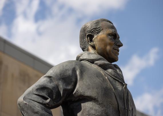 The head and shoulders of the Phog Allen statue against a blue sky and Allen Fieldhouse